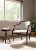 A classic armchair with a dark wood frame and textured light gray upholstery, styled in a sunlit room next to a small wooden side table and a potted plant.