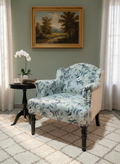 A classic upholstered armchair with a blue and white floral pattern, set in a styled room next to a small black side table with a white orchid, featuring a framed landscape painting on a sage green wall.