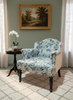 A classic upholstered armchair with a blue and white floral pattern, set in a styled room next to a small black side table with a white orchid, featuring a framed landscape painting on a sage green wall.