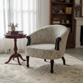 A classic upholstered armchair with a beige and gray floral pattern, dark wood arms and legs, styled in a cozy living room next to a small wooden side table and on a patterned area rug.