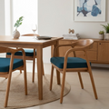 A modern dining room scene featuring a light oak wood dining table and four matching armchairs with teal blue upholstered seats, arranged on a round jute rug. A wooden sideboard, a framed abstract print, and a window are visible in the background.