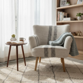 A cozy living room corner featuring a light beige armchair with a gray striped knit throw blanket draped over it, next to a small wooden side table with a mug and book. A light wood bookshelf stands in the background, and a patterned cream shag rug covers the floor.