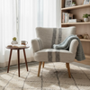 A cozy living room corner featuring a light beige armchair with a gray striped knit throw blanket draped over it, next to a small wooden side table with a mug and book. A light wood bookshelf stands in the background, and a patterned cream shag rug covers the floor.