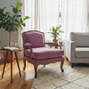 A styled living room scene featuring a dusty purple velvet armchair as the main product, set on a cream geometric pattern rug next to a light gray sofa, with a potted fiddle-leaf fig and other green plants in a bright room with sheer white curtains.
