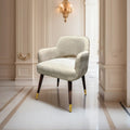 A light beige upholstered armchair with dark wood legs and gold metal tips, placed in an elegant cream-colored hallway with a gold wall sconce.