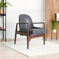 A modern gray upholstered armchair with dark wood arms and legs, placed on a white rug in a bright, minimalist living room with a slatted wood accent wall, plants, and a knit pouf in the foreground.