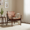 A mid-century modern armchair with a woven tan leather seat and backrest, styled in a bright living room next to a small wooden side table with a plant and mug.