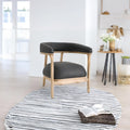A modern round area rug with black and white stripes and fringe trim, styled in a bright, minimalist room with a wooden-framed armchair and a small side table.