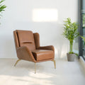 A modern brown leather armchair with gold-toned metal legs sits in a bright, minimalist room with white walls and light wood flooring. Several potted green plants are arranged around the space, and large floor-to-ceiling windows on the right offer a view of a city skyline.