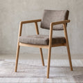 A modern wooden armchair with a light oak frame and taupe-gray velvet upholstery, set against a neutral concrete-textured wall and light stone floor.