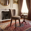 A classic interior room scene featuring a beige upholstered armchair on a red and blue patterned oriental rug, next to a small wooden side table, with a white fireplace, gold-framed mirror, and arched window with dusty rose curtains.