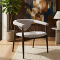 A modern upholstered accent chair with a curved backrest and dark wood legs, styled in a warm, contemporary living room setting with a potted plant, wall art, and a side table.