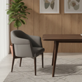 A modern dining room scene featuring a gray upholstered armchair next to a dark wood dining table on a patterned rug, with a potted plant and two framed abstract art prints on a wood-paneled wall.
