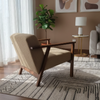 A modern mid-century style armchair with beige upholstery and dark wood legs, positioned on a patterned area rug in a styled living room with a gray sofa, side table, lamp, and abstract wall art.