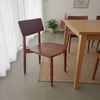 A mid-century modern wooden dining chair with a reddish-brown seat and backrest, positioned next to a light wood dining table on a woven jute rug in a bright room.