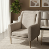 A modern high-back armchair upholstered in a textured beige bouclé fabric, set in a styled living room with a patterned rug, side table, lamp, and framed wall art.