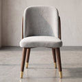 A modern accent chair with a light gray velvet upholstered seat and curved backrest, supported by dark wood legs with gold-tone metal caps, set against a textured neutral wall and concrete floor.
