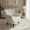 A cozy living room scene featuring a textured cream bouclé armchair in the foreground, with a light gray sofa, a patterned area rug, and two framed abstract prints on a warm beige wall.