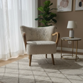 A styled living room corner featuring a cream bouclé armchair with wooden arms, set on a large cream and gray patterned area rug. The room has light wood floors, sheer white curtains, a small side table with a lamp, and abstract wall art.