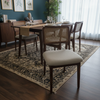 A dining room scene showing a wooden dining table with woven-back chairs and a patterned area rug in a styled interior with dark teal walls, framed botanical prints, pendant lights, and sheer curtains.
