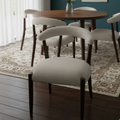 A dining room scene showing a round wooden table with six upholstered chairs in a light beige fabric, set on a patterned area rug against a dark teal wall with framed botanical prints.