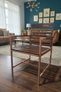 A wooden armchair with a slatted seat and backrest, shown in a furnished living room with wood floors, a rug, sofa, and wall decor.
