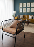 A modern black metal wire chair with a brown textured seat cushion in a styled living room with dark wood floors, a mustard yellow sofa, and a dark teal accent wall.