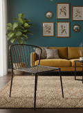 A modern black metal wire chair in the foreground of a styled living room with a mustard yellow sofa, teal wall, framed botanical prints, and a textured beige rug.