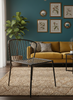 A modern black metal wire chair in the foreground of a styled living room with a mustard yellow sofa, teal wall, framed botanical prints, and a textured beige rug.