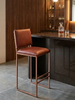 A modern bar stool with a brown leather seat and backrest, supported by a slim copper-toned metal frame, positioned in front of a dark wood bar counter in a stylish interior setting.