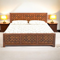 A dark brown wooden bed frame with intricate geometric lattice carvings on the headboard and footboard, styled with a white floral-patterned bedspread and two matching nightstands with lit lamps.